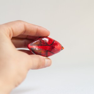 Peut inclure: Petit objet d&eacute;coratif rouge peint &agrave; la main, avec des touches de noir et de marron. L'objet est tenu dans la main d'une personne sur fond blanc. Il a une forme arrondie avec une extr&eacute;mit&eacute; pointue.