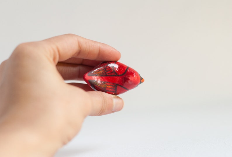 Peut inclure: Petit objet d&eacute;coratif rouge peint &agrave; la main, avec des touches de noir et de marron. L'objet est tenu dans la main d'une personne sur fond blanc. Il a une forme arrondie avec une extr&eacute;mit&eacute; pointue.