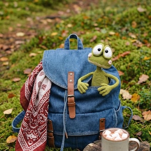 May include: A denim backpack with brown leather straps and a red and white patterned scarf. A green crocheted frog is peeking out of the front pocket. A mug of hot chocolate sits on a tree stump.