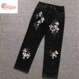 May include: Black denim jeans with decorative cross appliques in shades of gray and white. The jeans have a classic five-pocket design and a straight leg fit. The crosses are clustered on the front and back of the jeans.