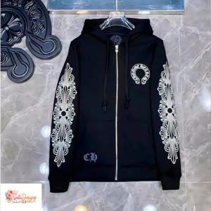 May include: Black zip-up hoodie with white ornate designs on the sleeves and a circular emblem on the chest. The hoodie features a silver zipper and the letters "CH" near the bottom. The hoodie is hanging on a hanger.