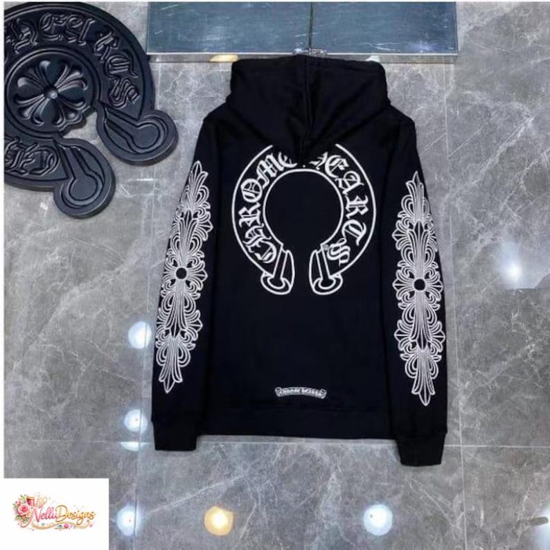 May include: Black hoodie with white ornate designs on the sleeves and back. The back features a large white emblem with the words "Chrome Hearts" in a circular design. The hoodie has a drawstring hood and a small logo near the bottom hem.
