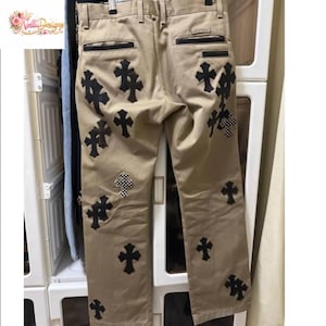 Pu&ograve; includere: Pantaloni color cachi con applicazioni a croce nere. Le croci sono di varie dimensioni e design, alcune con un motivo a scacchi. I pantaloni hanno un classico design a cinque tasche.