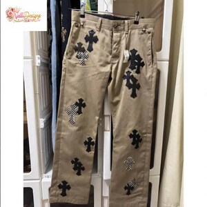 Pu&ograve; includere: Pantaloni color cachi con molteplici applicazioni di croci nere e croci a scacchi. I pantaloni hanno un classico design a cinque tasche e sono appesi a un'appendiabiti. Il marchio "Velli Designs" &egrave; visibile in alto a sinistra.