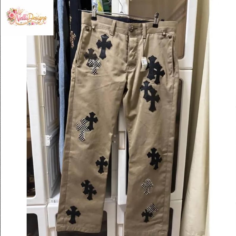 Pu&ograve; includere: Pantaloni color cachi con molteplici applicazioni di croci nere e croci a scacchi. I pantaloni hanno un classico design a cinque tasche e sono appesi a un'appendiabiti. Il marchio "Velli Designs" &egrave; visibile in alto a sinistra.