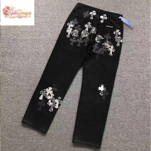 May include: Black denim jeans with multiple cross-shaped embellishments in silver and white. The jeans have a classic five-pocket design and a straight leg fit. The embellishments are clustered on the front and back of the jeans.
