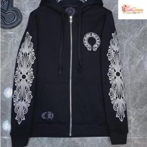 May include: Black zip-up hoodie with white ornate cross designs on the sleeves and a circular emblem on the chest. Features a silver zipper and the letters "CH" embroidered on the lower left. The hoodie has a hood and a small logo in the upper right corner.