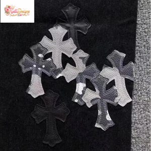 May include: A collection of decorative cross patches in various sizes and designs. The patches feature a mix of black, white, and gray colors with a textured appearance. Some crosses have a camouflage-like pattern.