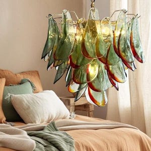 Pode incluir: Cena de quarto com cama laranja, almofadas e uma manta verde. Uma luminária decorativa com elementos de vidro verde e amarelo pende sobre a cama. O ambiente é acolhedor e convidativo.