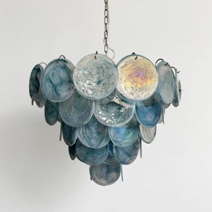 Peut inclure: Un lustre décoratif avec une cascade de disques ronds en verre bleu-vert translucide. Les disques sont suspendus à une chaîne argentée, créant un luminaire élégant et superposé. Le verre a un effet marbré.