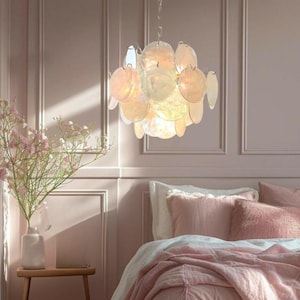 May include: A decorative chandelier with a cluster of iridescent, circular, shell-like elements. The light fixture hangs from a silver chain. The room features a pink colour scheme, with a bed and floral arrangement.