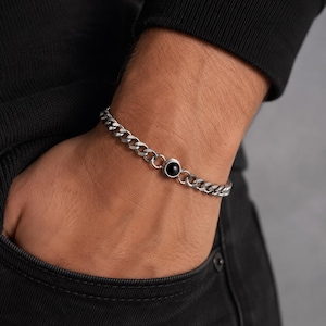 Peut inclure: Un bracelet à maillons gourmette de couleur argentée orné d'une pierre noire polie, circulaire et sertie dans une monture en métal. Le bracelet est composé de maillons plats entrelacés formant une chaîne continue avec un fermoir intégré.