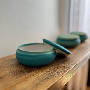 May include: A round ceramic container with a vibrant turquoise exterior and a smooth white interior. The matching turquoise lid is tilted and resting partially on the rim of the container, revealing the interior space and the thick ceramic walls.