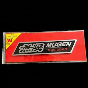 May include: A rectangular sticker on a red background. The sticker has a silver and black design with the word "MUGEN" and Japanese characters. The text "equipped" is also present. The sticker is packaged in a clear plastic sleeve.