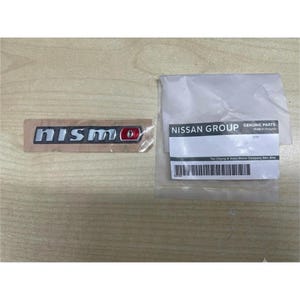 Könnte beinhalten: Ein Nismo-Emblem mit silberner Schrift und einem roten O, verpackt in durchsichtiger Schutzfolie. Daneben liegt ein weißer Plastikbeutel mit einem Nissan Group Genuine Parts-Etikett, einem Strichcode und dem Text Tan Chong & Sons Motor Company.