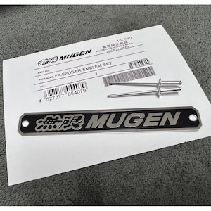 May include: A black and silver Mugen FR.Spoiler Emblem Set. The set features a black rectangular emblem with the Mugen logo and text, plus two silver mounting pins. A white paper with product details is also included.