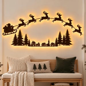 Puede incluir: Una pieza de arte mural de metal marrón oscuro con la silueta de Santa Claus en un trineo tirado por seis renos volando sobre un bosque de pinos y un pequeño pueblo. El diseño está retroiluminado con un brillo cálido y festivo.