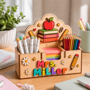 Op de afbeelding: Een houten bureau-organizer met de naam Mrs. Miller in kleurrijke 3D-letters. Het achterpaneel toont een rode appel, gestapelde boeken, linialen en een paperclip. De voorzijde bevat madeliefjes, een potloodafbeelding en een vakje.