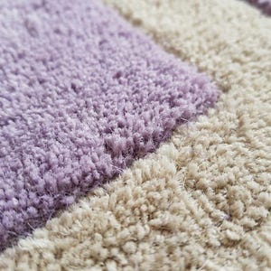 May include: Close-up of a soft, textured rug with a pattern of alternating lavender and beige rectangular blocks. The plush fibers create a tactile, inviting appearance, suitable for home decor.