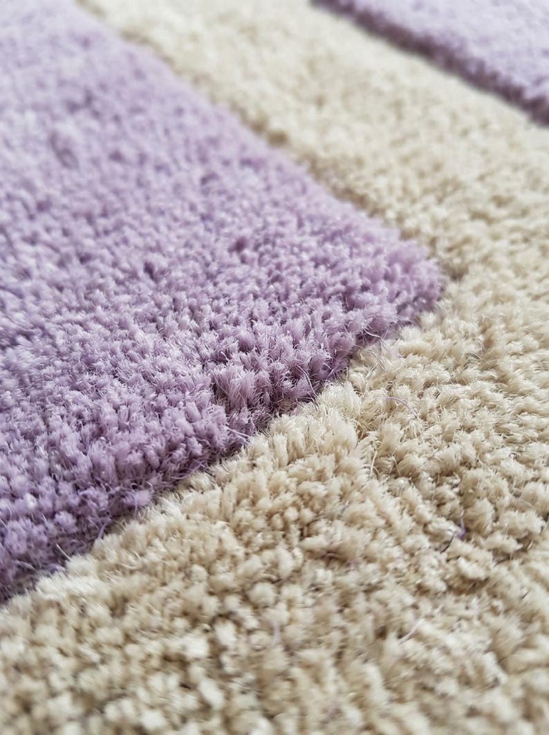 May include: Close-up of a soft, textured rug with a pattern of alternating lavender and beige rectangular blocks. The plush fibers create a tactile, inviting appearance, suitable for home decor.