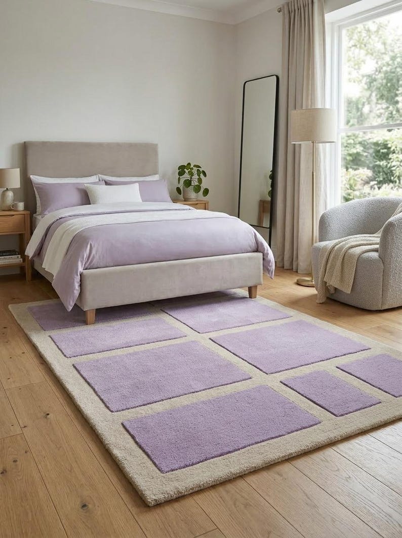 May include: A bedroom scene featuring a bed with lavender bedding and a matching area rug. The rug has a beige border with lavender rectangular inserts. A tall mirror and a cozy armchair complete the room's decor.