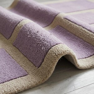 May include: A rolled-up, rectangular rug with a geometric pattern. The rug features a soft, textured pile in a light purple color, with a beige border and square accents. The rug is made of a plush material.