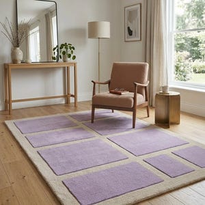 May include: A rectangular area rug with a light beige border and lavender-colored square inserts. The rug is placed on a hardwood floor in a room with a chair, side table, and mirror. The room has a neutral color palette.