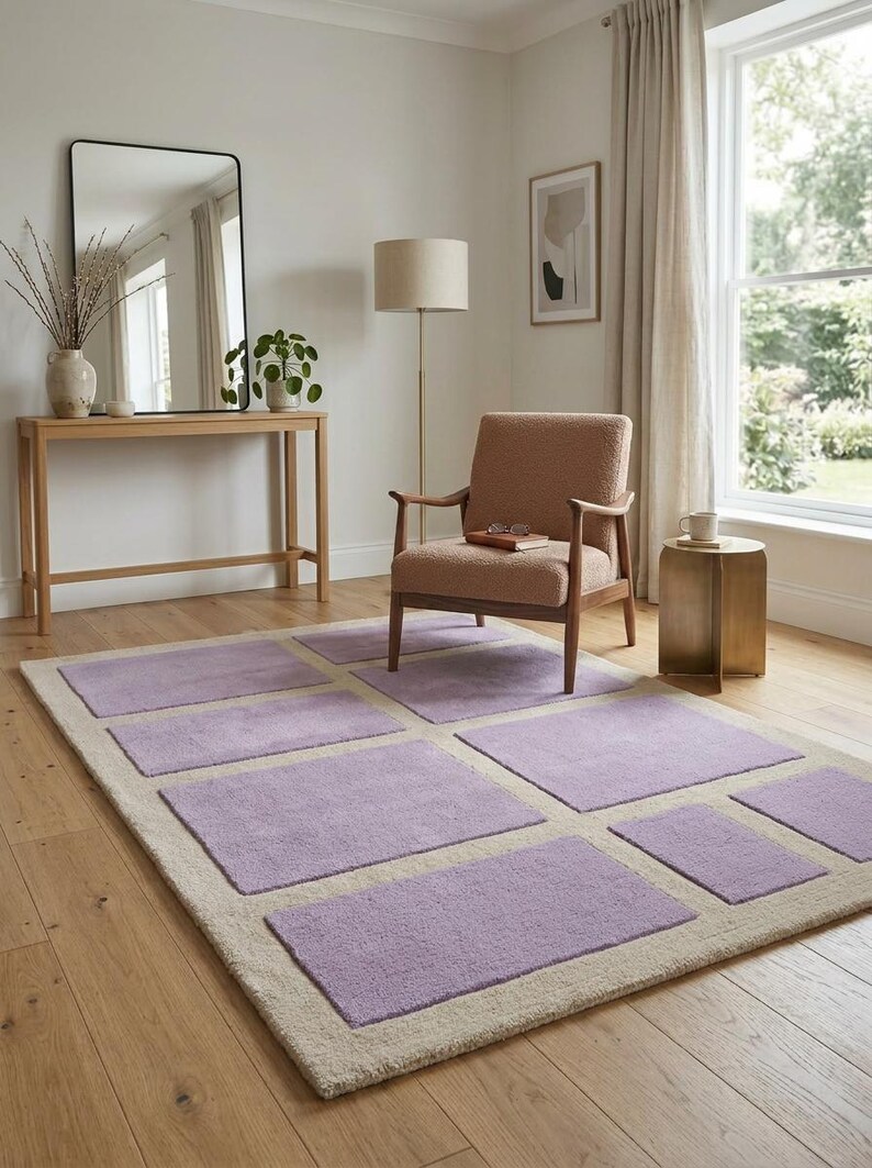 May include: A rectangular area rug with a light beige border and lavender-colored square inserts. The rug is placed on a hardwood floor in a room with a chair, side table, and mirror. The room has a neutral color palette.