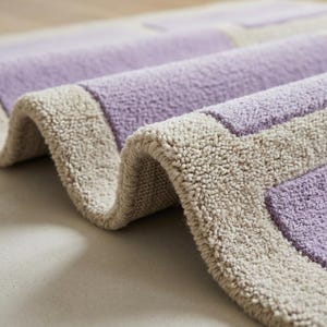 May include: Close-up of a rolled-up, rectangular bath mat. The mat features a light purple center with a beige border. The texture appears soft and plush, with a slight curve visible in the rolled section.