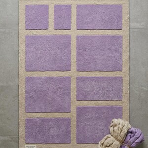 May include: A rectangular bath mat with a light beige border and several lilac-colored rectangular inserts. A bundle of beige and lilac yarn is in the lower right corner. The mat is on a gray tiled floor.