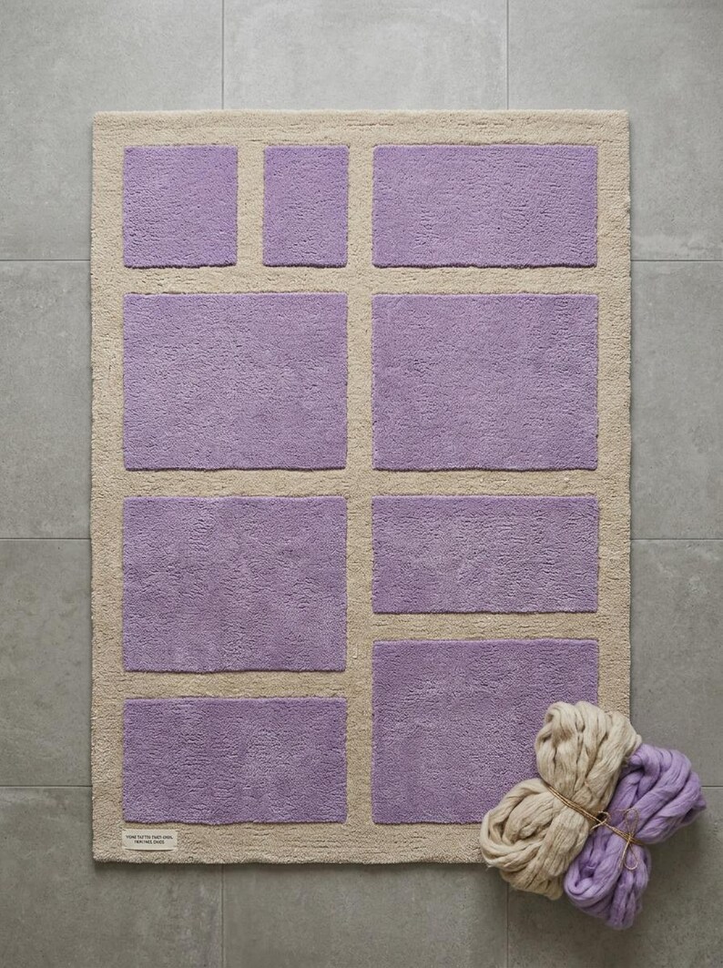 May include: A rectangular bath mat with a light beige border and several lilac-colored rectangular inserts. A bundle of beige and lilac yarn is in the lower right corner. The mat is on a gray tiled floor.