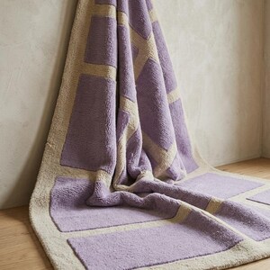 May include: A rectangular bath mat with a geometric design. The mat features a light purple color with beige rectangular accents and border. The mat is made of a soft, plush material and is displayed on a wooden floor.