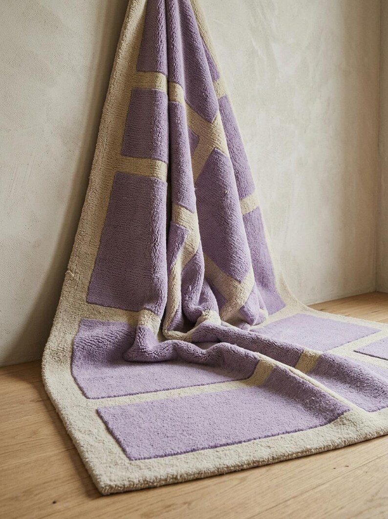 May include: A rectangular bath mat with a geometric design. The mat features a light purple color with beige rectangular accents and border. The mat is made of a soft, plush material and is displayed on a wooden floor.