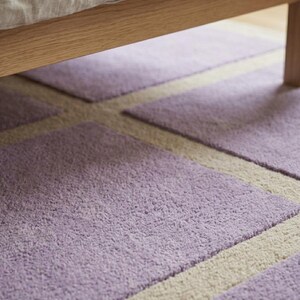 May include: Close-up of a lavender and cream-colored rug with a geometric pattern. The rug is under a wooden bed frame. The rug's texture is soft and plush, with a short pile. The design features rectangular shapes.