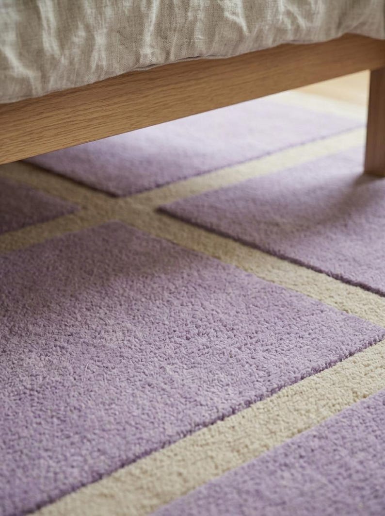 May include: Close-up of a lavender and cream-colored rug with a geometric pattern. The rug is under a wooden bed frame. The rug's texture is soft and plush, with a short pile. The design features rectangular shapes.