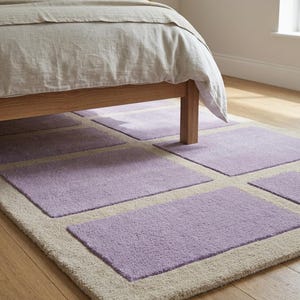 May include: A rectangular area rug with a light beige border and lavender-colored square inserts. The rug is placed on a wooden floor, partially under a bed with a light-colored comforter. The rug's dimensions are not visible.