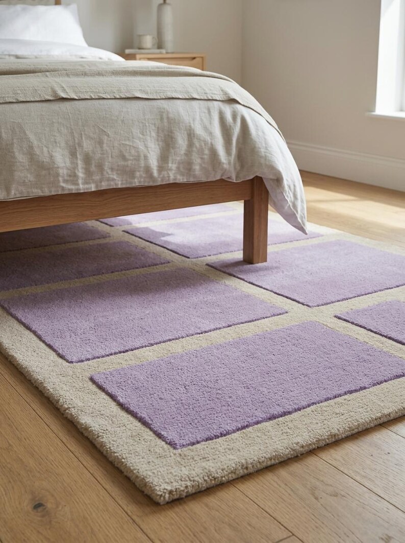 May include: A rectangular area rug with a light beige border and lavender-colored square inserts. The rug is placed on a wooden floor, partially under a bed with a light-colored comforter. The rug's dimensions are not visible.