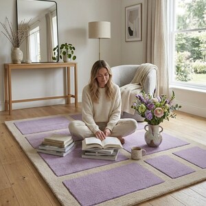 May include: A rectangular area rug with a light purple and beige geometric design. The rug is in a living room setting with a person sitting on it, surrounded by books and a vase of flowers. The rug's dimensions are approximately 6 x 8 feet.