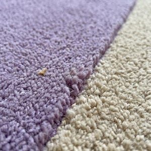May include: Close-up of a section of a rug with a textured surface. The rug features a color block design with a light purple section and a beige section. The fibers are visible, creating a soft, plush appearance.