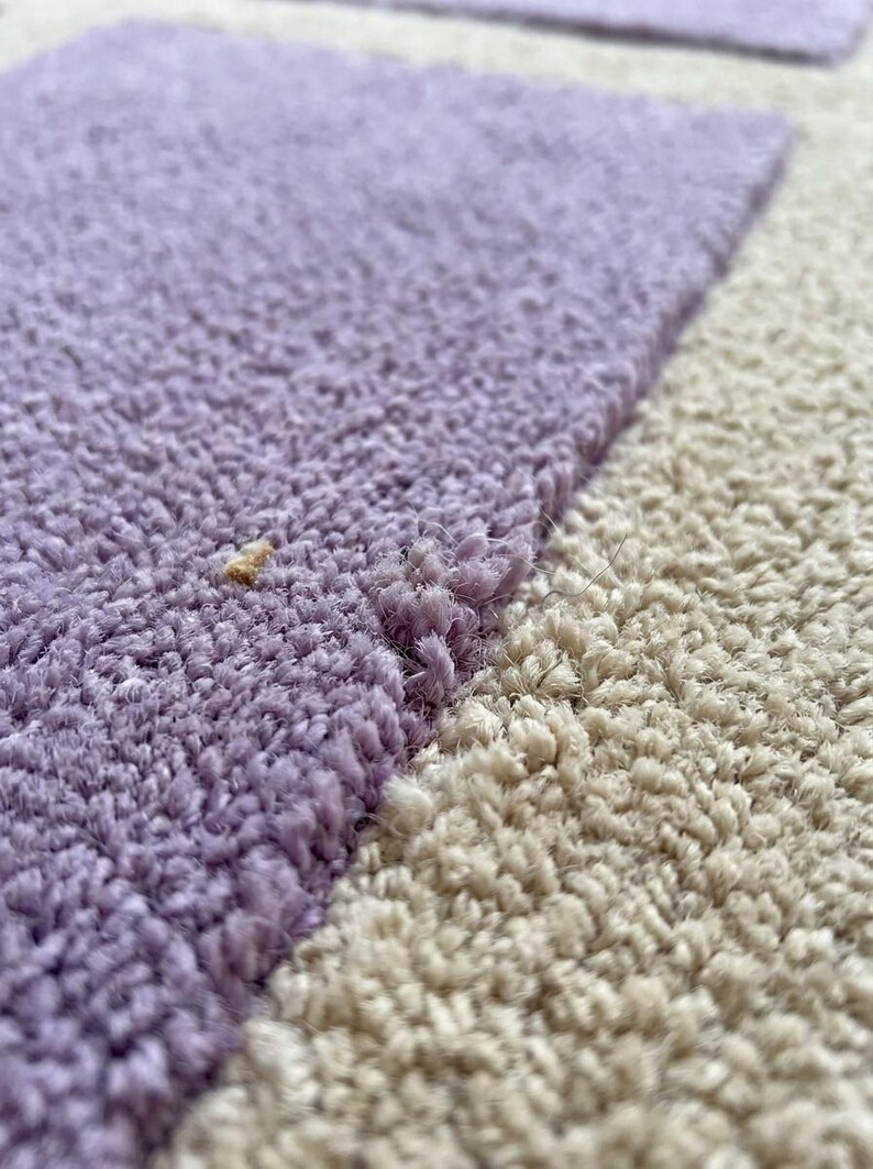 May include: Close-up of a section of a rug with a textured surface. The rug features a color block design with a light purple section and a beige section. The fibers are visible, creating a soft, plush appearance.