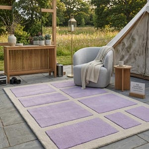 May include: A rectangular rug with a cream-colored border and lavender-colored square accents. The rug is placed on a stone patio, with a chair, side table, and tent in the background. The rug is part of "The Lavender Collection".
