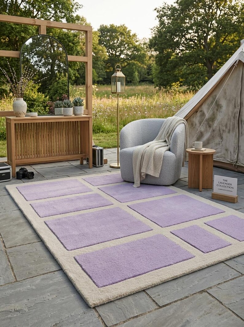 May include: A rectangular rug with a cream-colored border and lavender-colored square accents. The rug is placed on a stone patio, with a chair, side table, and tent in the background. The rug is part of "The Lavender Collection".