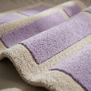May include: A close-up of a soft, plush bath mat with a geometric design. The mat features alternating stripes of light beige and lavender. The texture appears to be absorbent and comfortable, suitable for a bathroom setting.