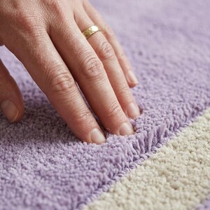 May include: Close-up of a soft, textured rug with a lavender and off-white striped pattern. The rug's fibers are visible, and a hand with a gold ring is touching the surface, highlighting the texture and color variations.