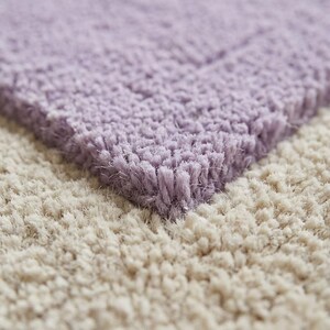 May include: Close-up of a soft, textured rug with a two-tone design. The top section is a light lavender color, while the bottom section is a creamy beige. The rug has a plush, shaggy appearance, suggesting comfort and warmth. Ideal for home decor.