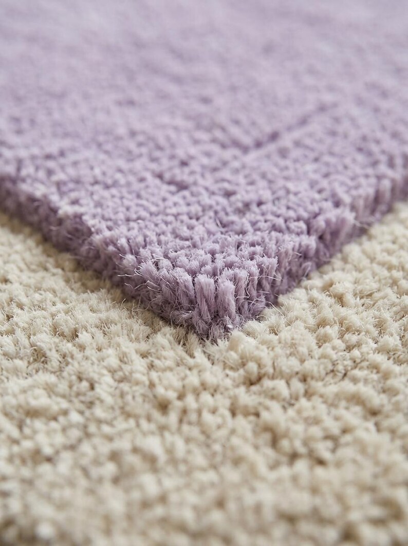 May include: Close-up of a soft, textured rug with a two-tone design. The top section is a light lavender color, while the bottom section is a creamy beige. The rug has a plush, shaggy appearance, suggesting comfort and warmth. Ideal for home decor.