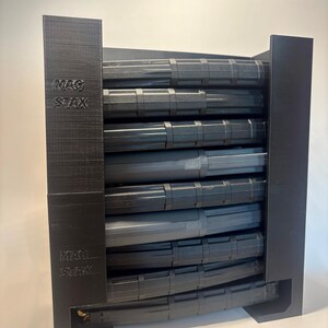 May include: A black, 3D-printed magazine rack with the text "MAG STAX" on the side. The rack holds several black magazines, visible from the side. The rack is designed to hold magazines in a vertical orientation.