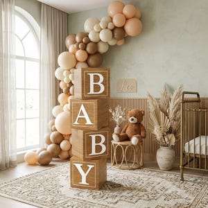 Puede incluir: Una escena de guardería con un arco de globos en tonos beige y marrón, bloques de madera que deletrean "BABY" y un oso de peluche. La habitación cuenta con una alfombra, una cuna y hierba de la pampa decorativa, creando un ambiente cálido.