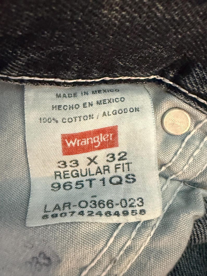 K&ouml;nnte beinhalten: Nahaufnahme eines Wrangler Jeansetiketts. Das Etikett zeigt "MADE IN MEXICO", "HECHO EN MEXICO" und "100% COTTON / ALGODON". Die Gr&ouml;&szlig;e ist 33 x 32, Regular Fit, mit einem Produktcode.