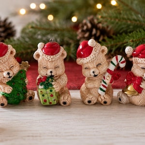 Puede incluir: Cuatro adornos de cerámica con forma de oso, cada uno con un gorro de Papá Noel rojo con borde blanco. Los osos son de color marrón claro con pelaje texturizado. Sostienen un árbol de Navidad verde, un regalo verde, un bastón de caramelo y una campana.